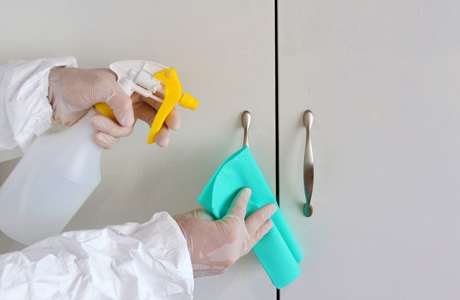 Gloved hands cleaning cabinet handles with a spray bottle and cloth for hygiene.